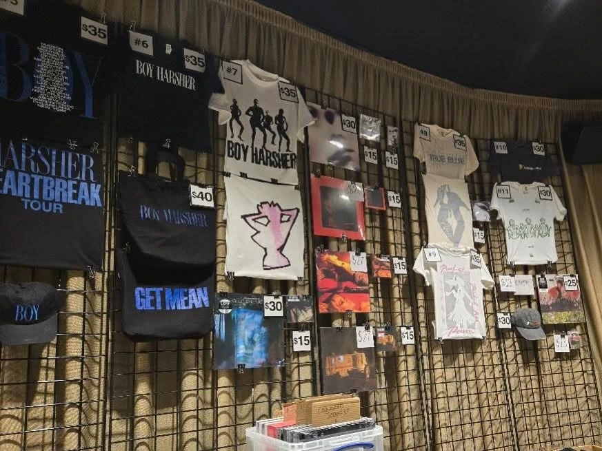 Boy Harsher merchandise wall with numbered shirts, a blue-lettered tote and music releases at Hollywood Palladium