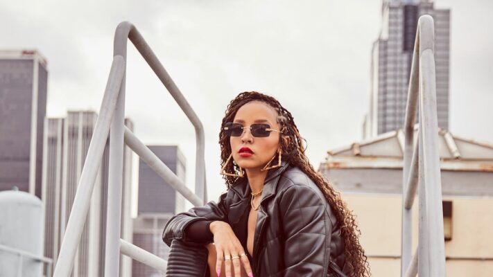 Tinashe in a black quilted coat, knee-high boots and sunglasses in a rooftop fashion portrait