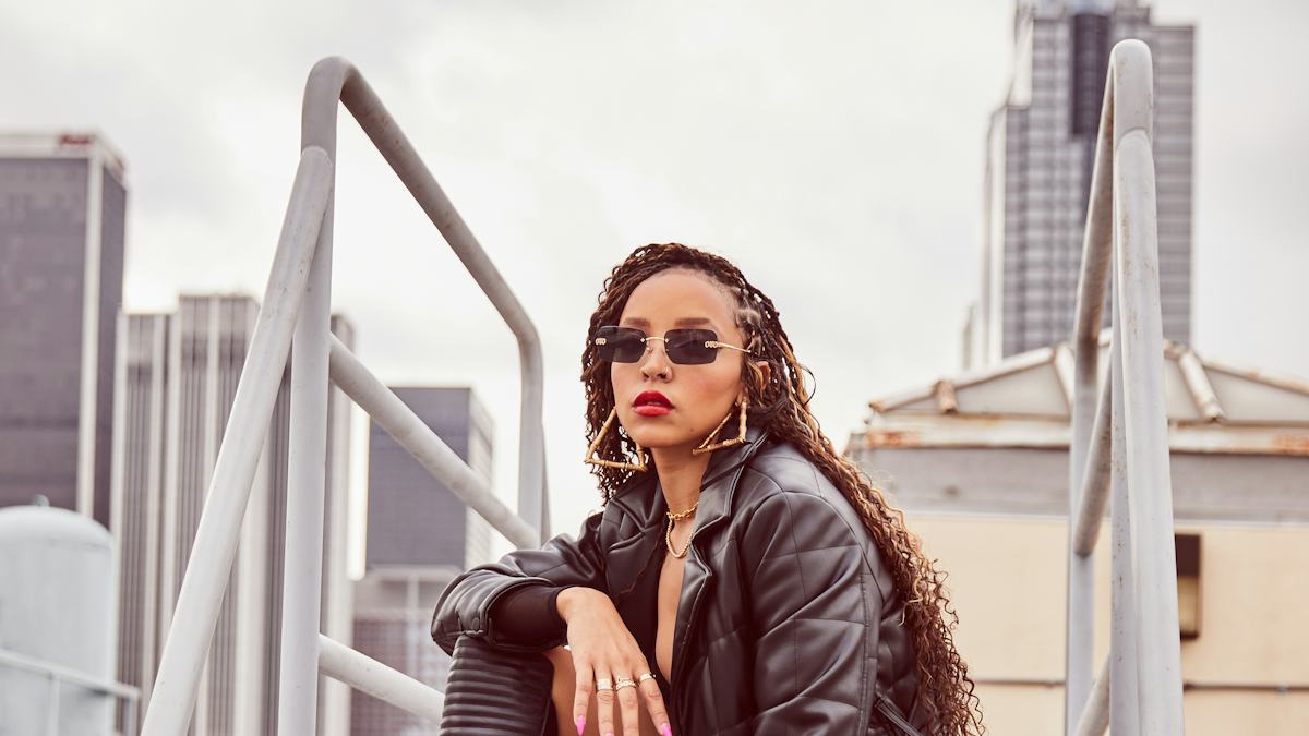 Tinashe in a black quilted coat, knee-high boots and sunglasses in a rooftop fashion portrait